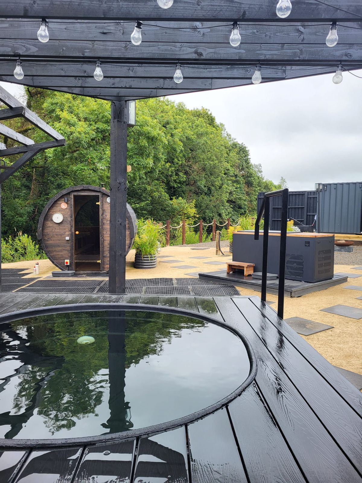 Looking across the round pool on the dark timber deck to the barrel sauna, under the pergola lights