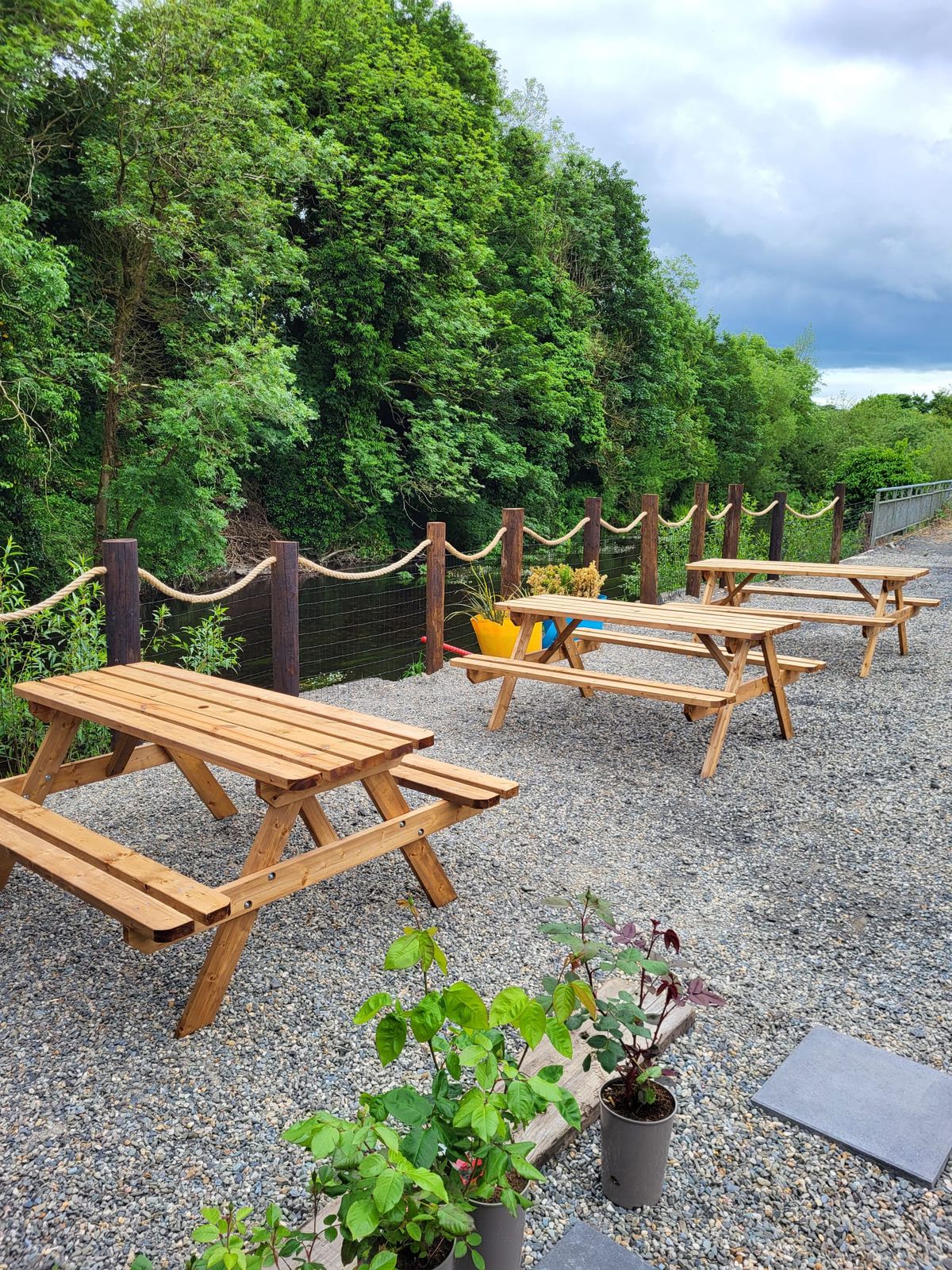 Picnic tables on the gravel beside the rope fence and the riverbank trees