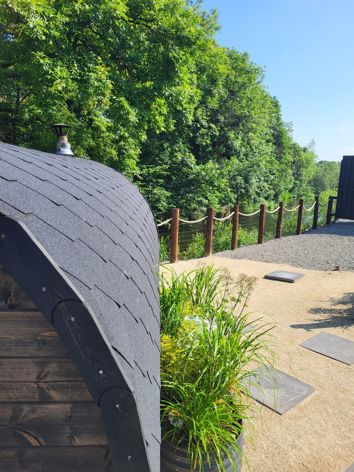 The shingled roof of the barrel sauna beside the rope-fenced path along the river
