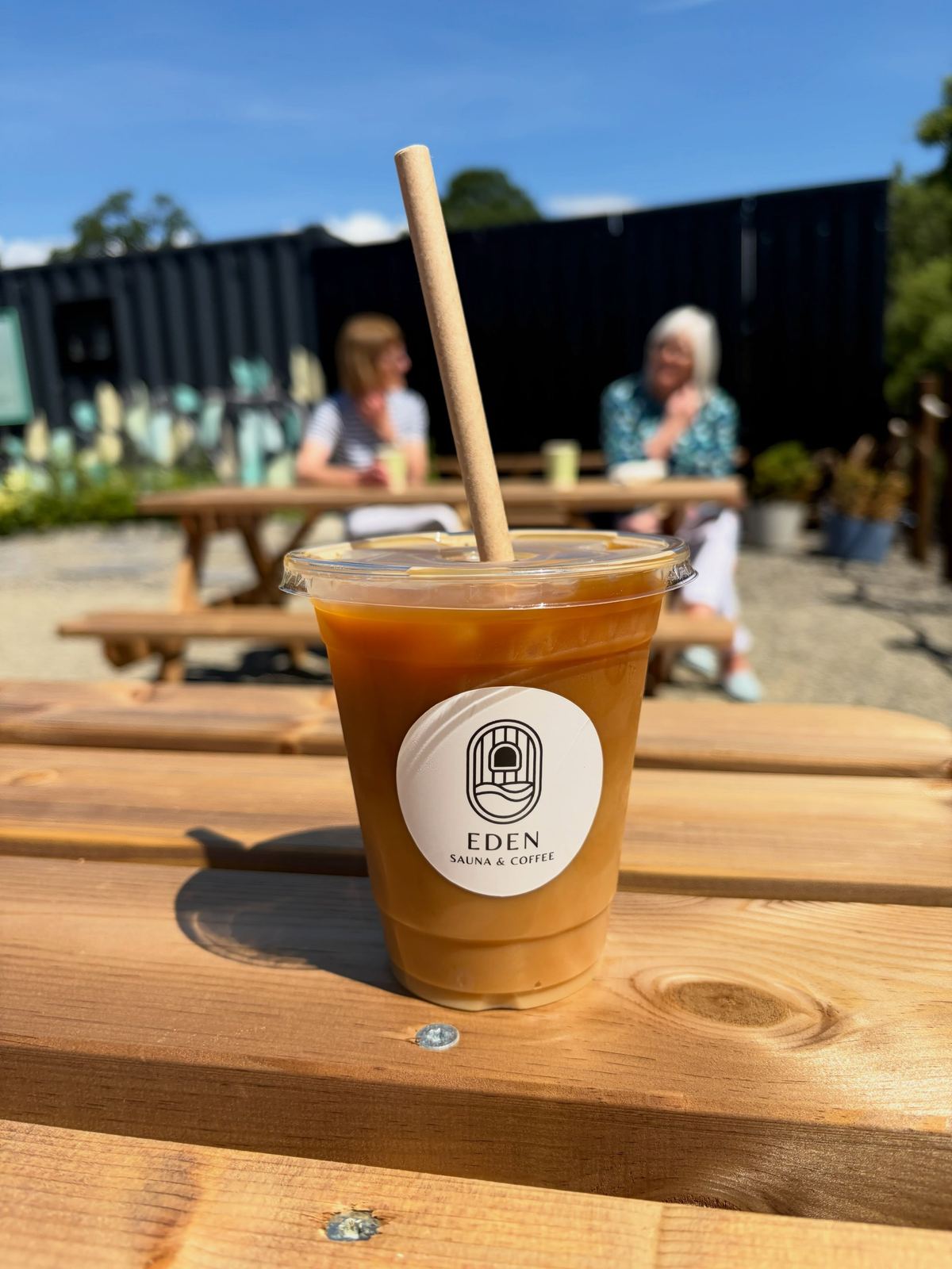 An iced latte on a picnic table, with people chatting in the sun behind
