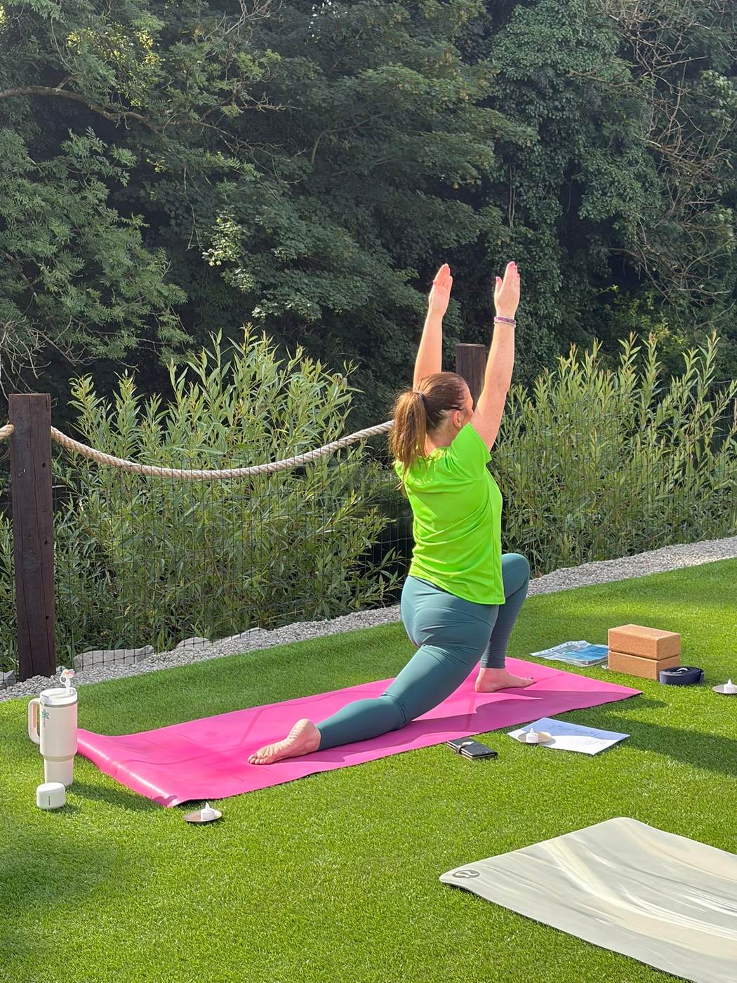 A yoga teacher in a low lunge on the lawn, with the riverbank trees behind